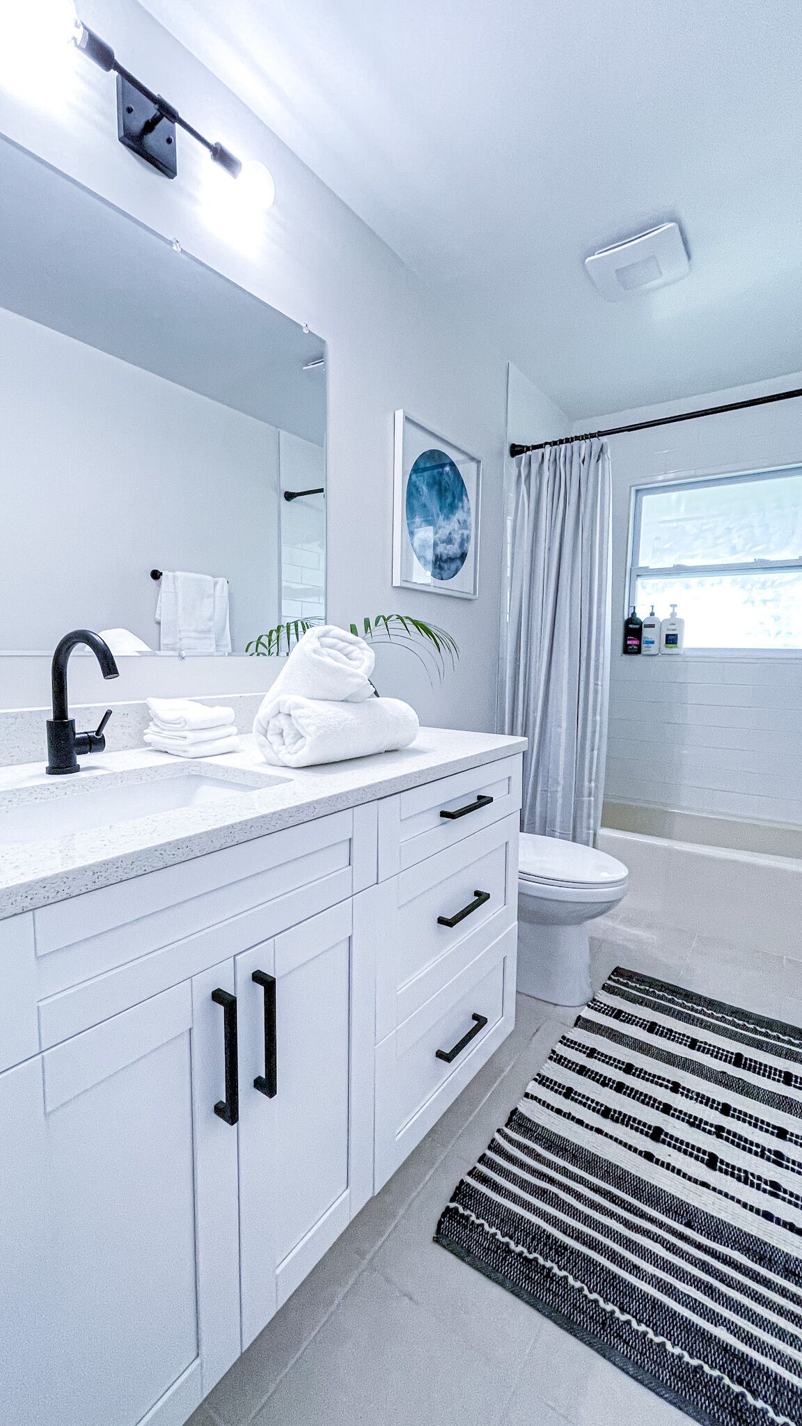 Un baño moderno con tocador blanco, grifo negro y un espejo grande. Hay un inodoro y una ventana con cortinas.
