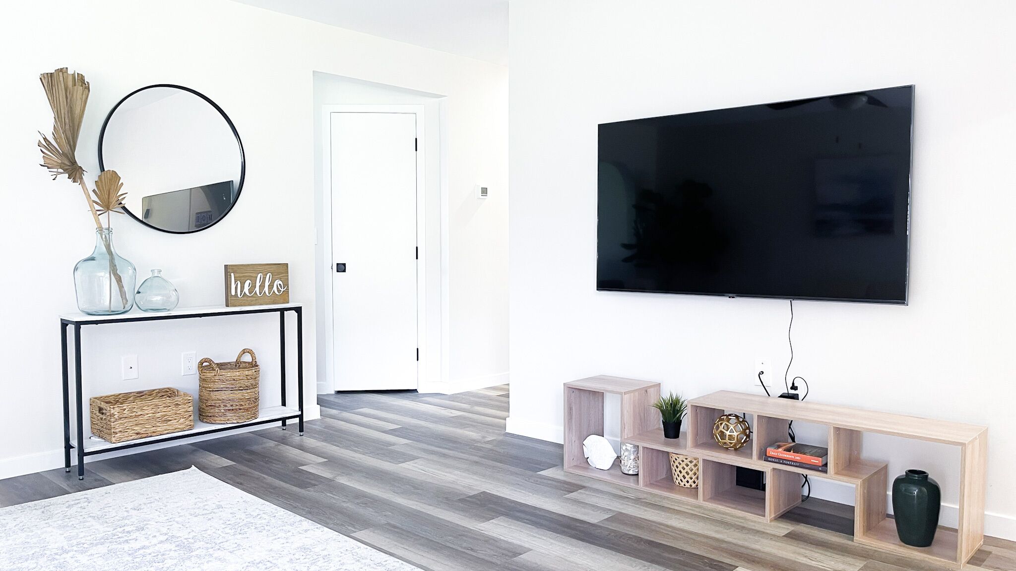 Un soggiorno moderno con una TV a schermo piatto montata a parete, un tavolino consolle con cesti, uno specchio e una porta che dà su un'altra stanza.