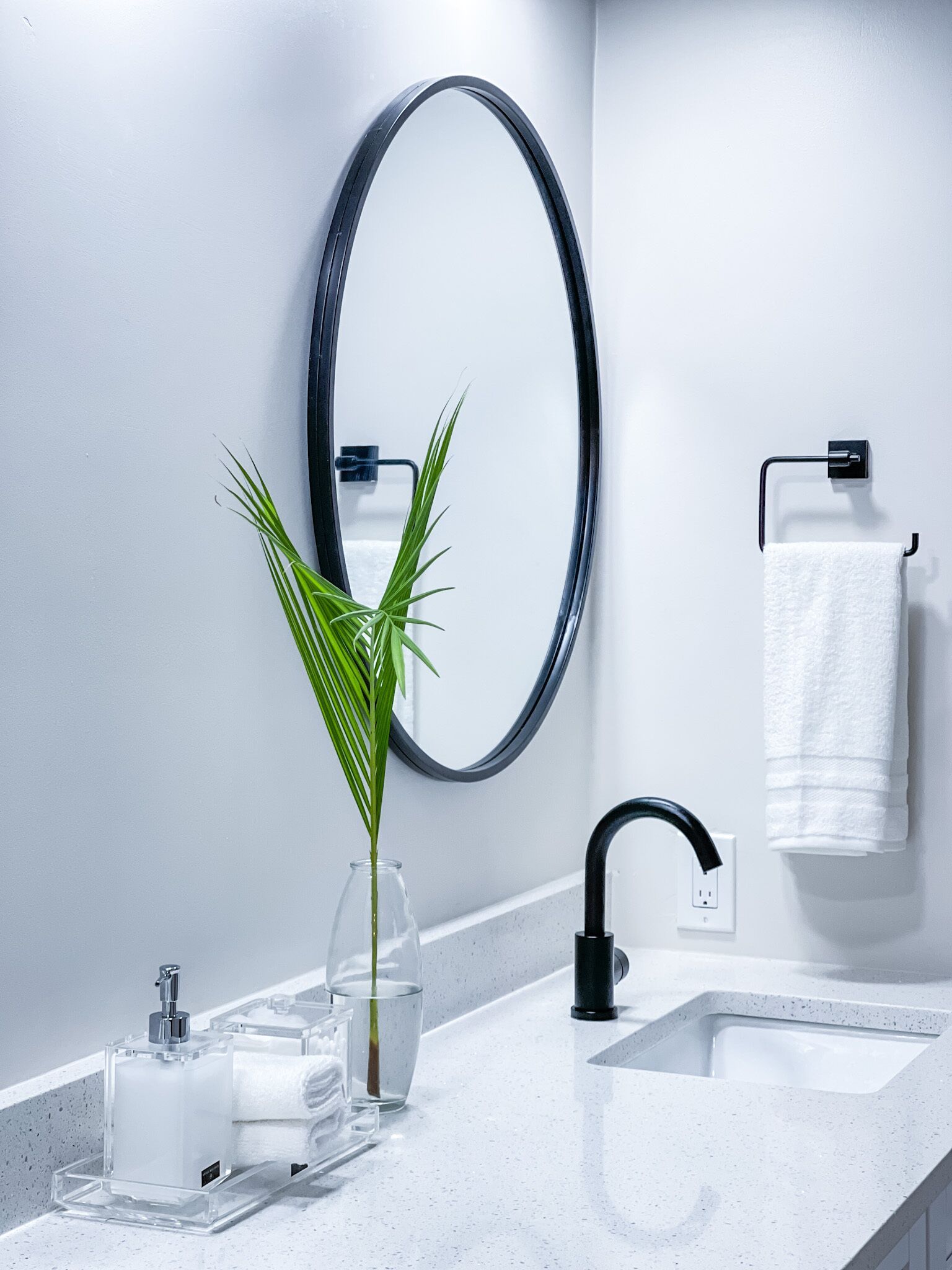 Ein modernes Badezimmer mit weißer Ablagefläche, einem schwarzen Wasserhahn, einer Glasvase mit Pflanze, einem Seifenspender und einem Handtuch an einem Haken.
