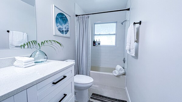 A modern bathroom with a white vanity, a glass vase with a plant, a shower with a glass door, and a window providing natural light. - Relaxing 2 BED home near Manasota Beach. (Venice)