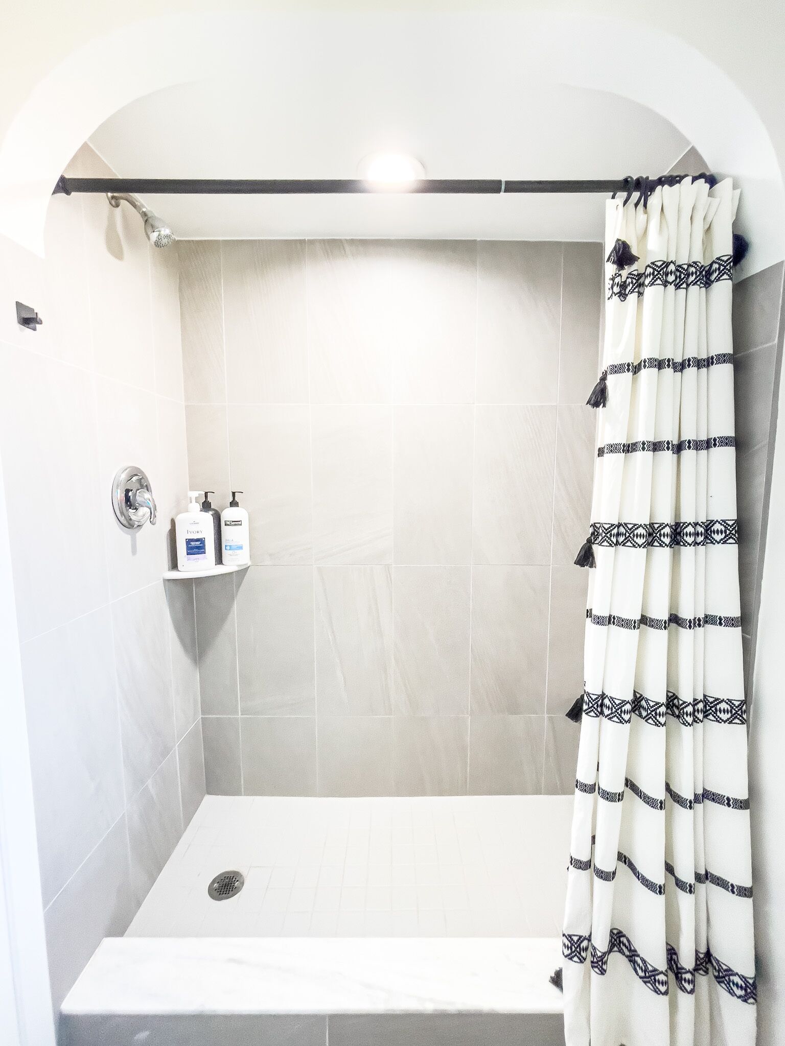 Uma área de chuveiro moderna com parede revestida de azulejos brancos, cortina de chuveiro com padrão geométrico e duas garrafas de shampoo em uma prateleira.