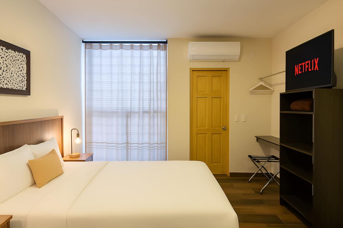 A hotel room with a bed, a wardrobe, a television displaying 'NETFLIX', a window with curtains, and an air conditioning unit.