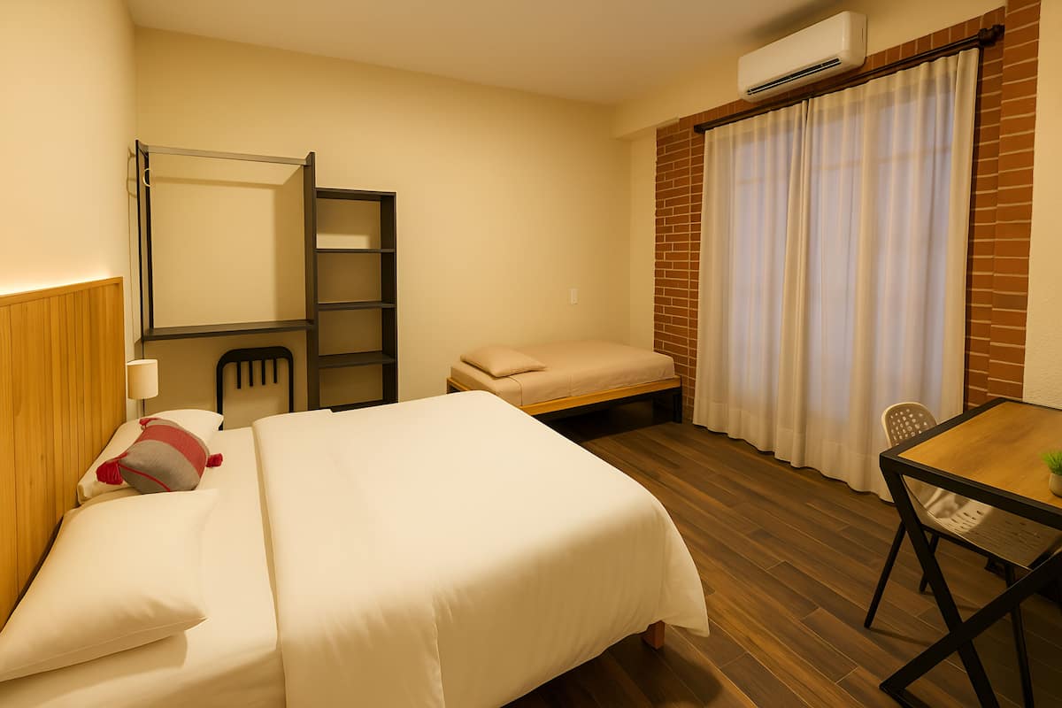 A hotel room with a bed, a desk, a chair, a small table, a lamp, a window with curtains, and an air conditioning unit.