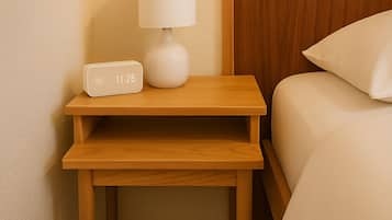 Una mesita de noche de madera bien ordenada con un reloj despertador blanco y una lámpara, ubicada junto a una cama con un almohadón blanco.