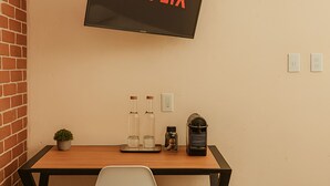 A room with a TV mounted on the wall displaying "NETFLIX", a wooden desk with a chair, two bottles, a coffee maker, and a small potted plant. - Casa Acueducto (Oaxaca)