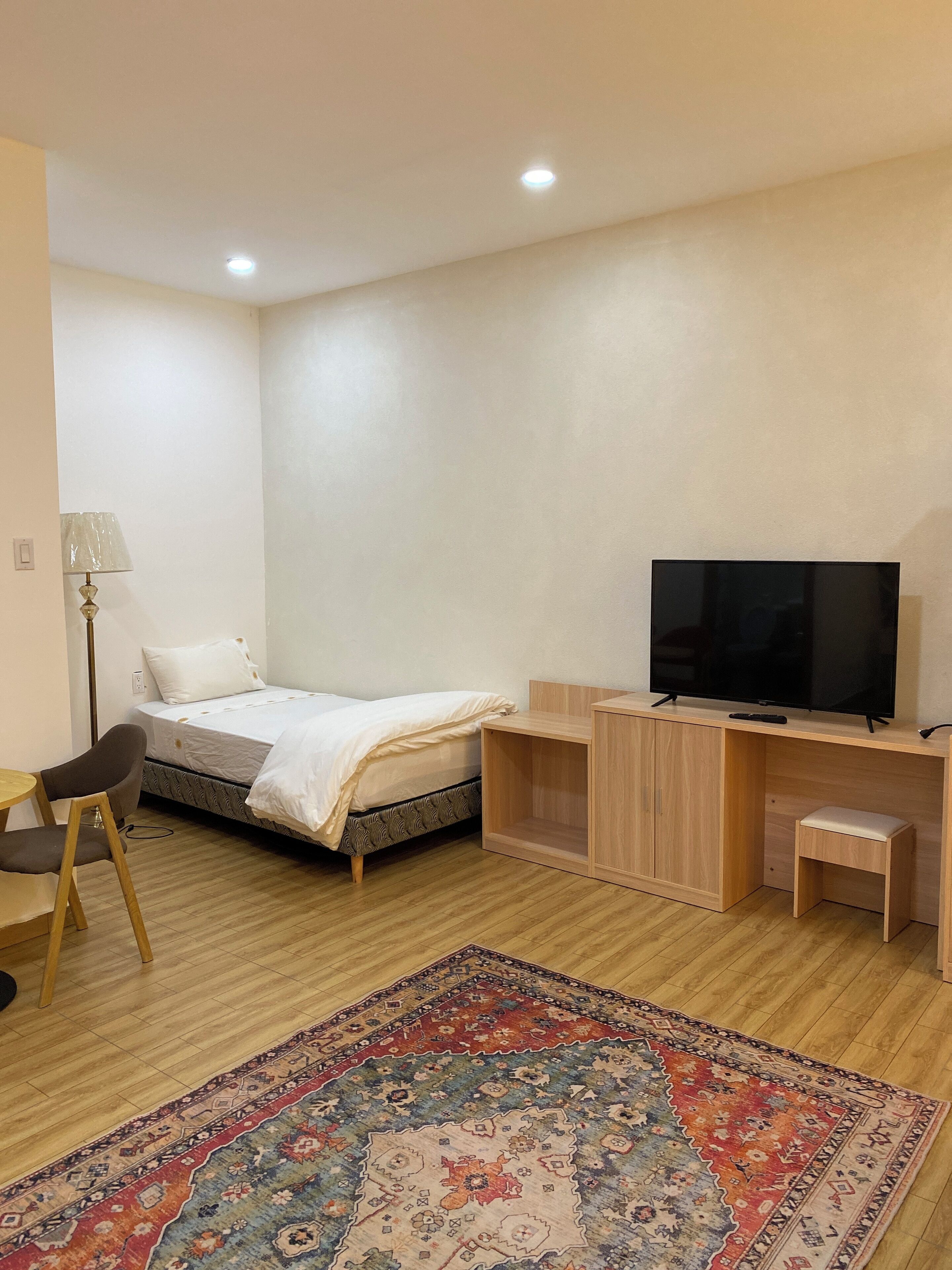 Un dormitorio con cama, televisor, escritorio, silla, taburete pequeño y alfombra estampada.