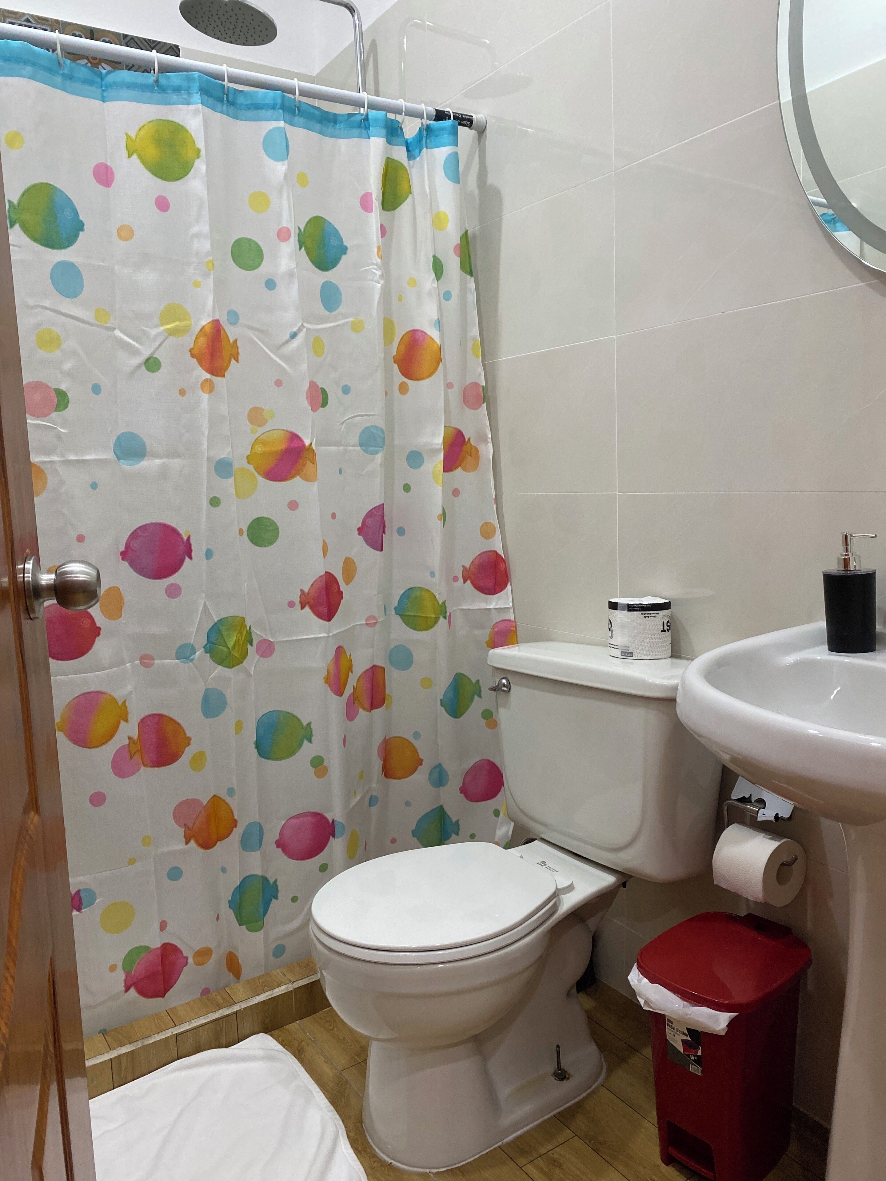 Un baño con inodoro, lavabo, un cubo de basura rojo y cortina de ducha con estampado de peces de colores.