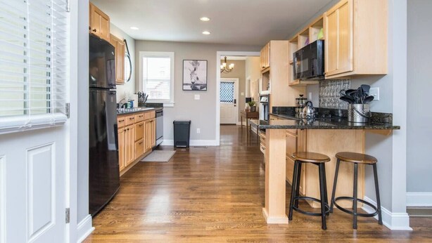 A modern kitchen with wooden cabinets, a black refrigerator, and a granite countertop. There are two stools and a trash can visible. - Location, Location, Location (Lexington)