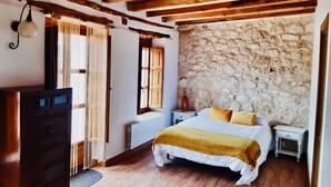 A bedroom with a stone wall, wooden ceiling beams, a bed with white bedding and a mustard throw, a nightstand, and a dresser. - Charming accommodation, next to Ribera del Duero and Hoces del Duratón (aldeasoña)