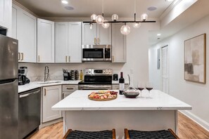Une cuisine moderne avec des meubles blancs, des appareils en acier inoxydable, un îlot central agrémenté de fruits et de bouteilles de vin, et des luminaires suspendus.