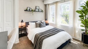 Un dormitorio con una cama, mesitas de noche, un estante en la pared con fotografías, una planta en maceta y una ventana con cortinas.
