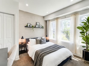 Un dormitorio con una cama, mesitas de noche, un estante en la pared con fotografías, una planta en maceta y una ventana con cortinas.
