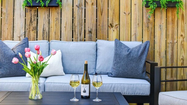 Ein Holzzaun, ein Sofa mit Kissen, ein Tisch mit Flasche und Gläsern sowie eine Vase mit Blumen.