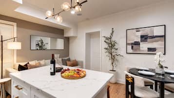 A modern kitchen with a marble island, a dining area with a black table and chairs, and a living area with a sofa and a plant.