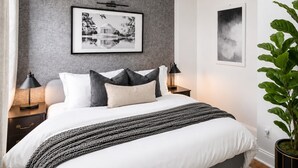 Ein Schlafzimmer mit einem Bett, zwei Nachttischen, einer Stehlampe, einer Topfpflanze und zwei gerahmten Bildern an der Wand.