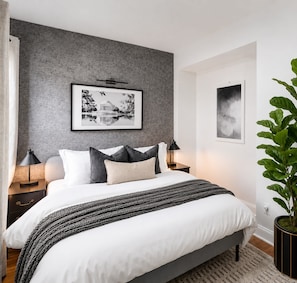 Un dormitorio con una cama, dos mesitas de noche, una lámpara auxiliar, una planta en maceta y dos cuadros en la pared.