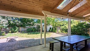 A covered patio area with a wooden table and benches, a fire pit, and a view of a backyard with a wooden fence and greenery. - Arioso House - Style Inspired by Classical Music! (Newberg)