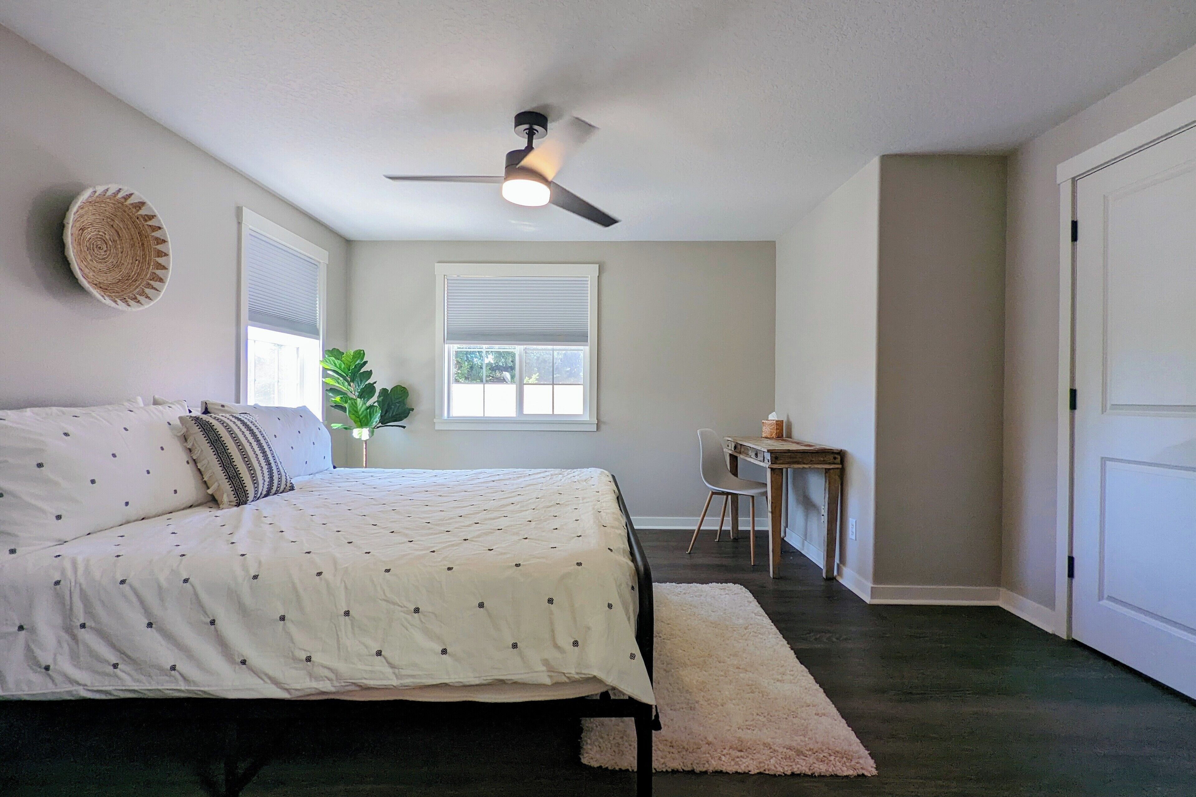 A bedroom with a bed, a desk, a chair, a ceiling fan, and two windows.