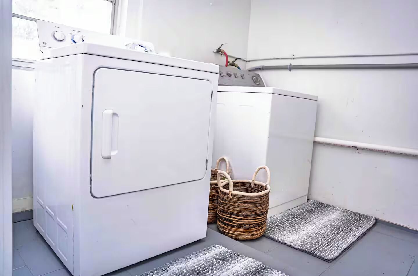 A laundry room with two white washing machines, wicker baskets, and grey textured mats on the floor.