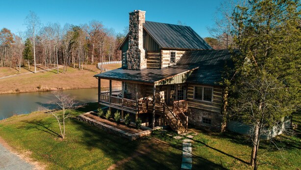 A log cabin with a porch, a stone chimney, and a pond in the background. - Rustic Luxury Near Charlottesville/Montpelier/Wineries /A Historic Cabin Retreat (Somerset)