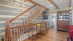 A wooden staircase with a railing, a dining area with chairs and a vase of flowers, and a wooden cabinet with glass doors. - Rustic Luxury Near Charlottesville/Montpelier/Wineries /A Historic Cabin Retreat (Somerset)