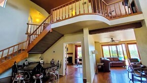 Amplia sala de estar con una escalera de madera curvada, un comedor con sillas de metal y un área de descanso con sofás y sillones.