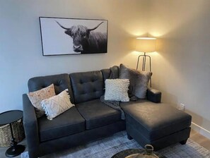 Un canapé d’angle gris foncé agrémenté de plusieurs coussins, un lampadaire noir et, au mur, une œuvre d’art représentant un taureau, encadrée.