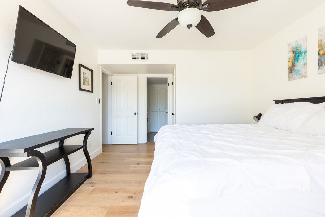 A bedroom with a bed, a desk, a ceiling fan, and a TV mounted on the wall.