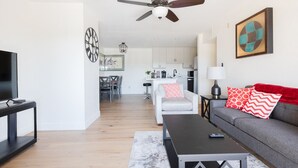 Un soggiorno moderno con un divano, un tavolino da caffè e una TV. Si nota una ventola a soffitto, una cucina sullo sfondo e opere d'arte alle pareti.