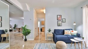 A modern living room with a blue sofa, a round ottoman, and a small side table with a lamp. There are two potted plants, one on the sofa and one on the side table. The room has light blue walls and a striped rug.