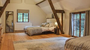 Une chambre avec un plafond en pente, des poutres en bois, une grande fenêtre, un lit, une table de chevet avec un vase et un radiateur.