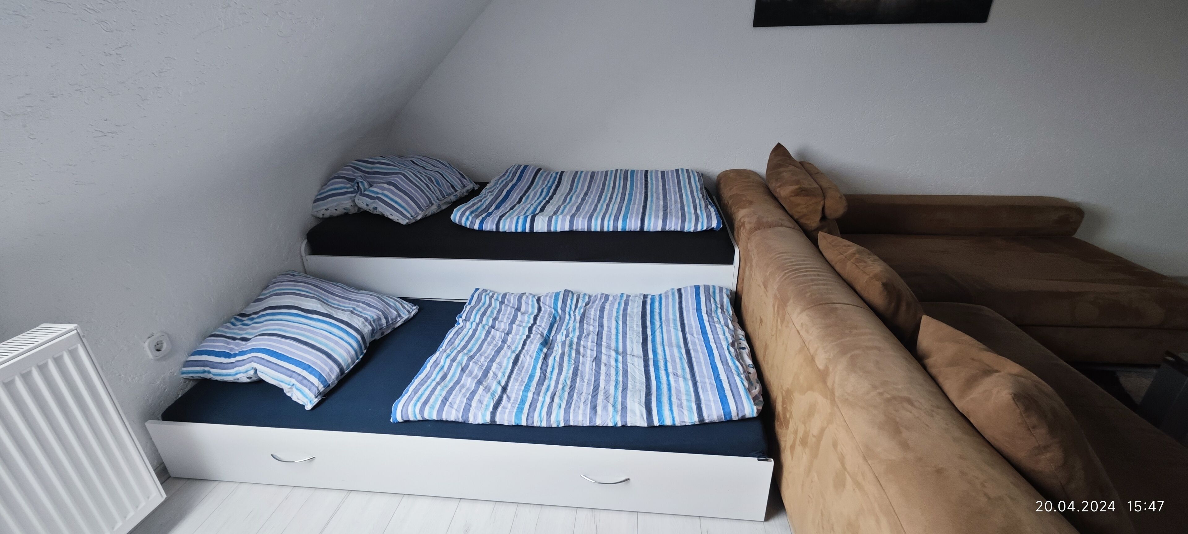 A bedroom with two beds, each with striped bedding and pillows, a brown sofa, and a white radiator.