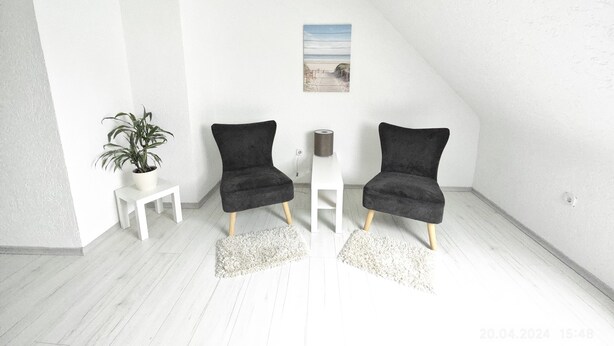 Una habitación minimalista con dos sillas negras, una mesita blanca, una planta en maceta y un cuadro con una playa.