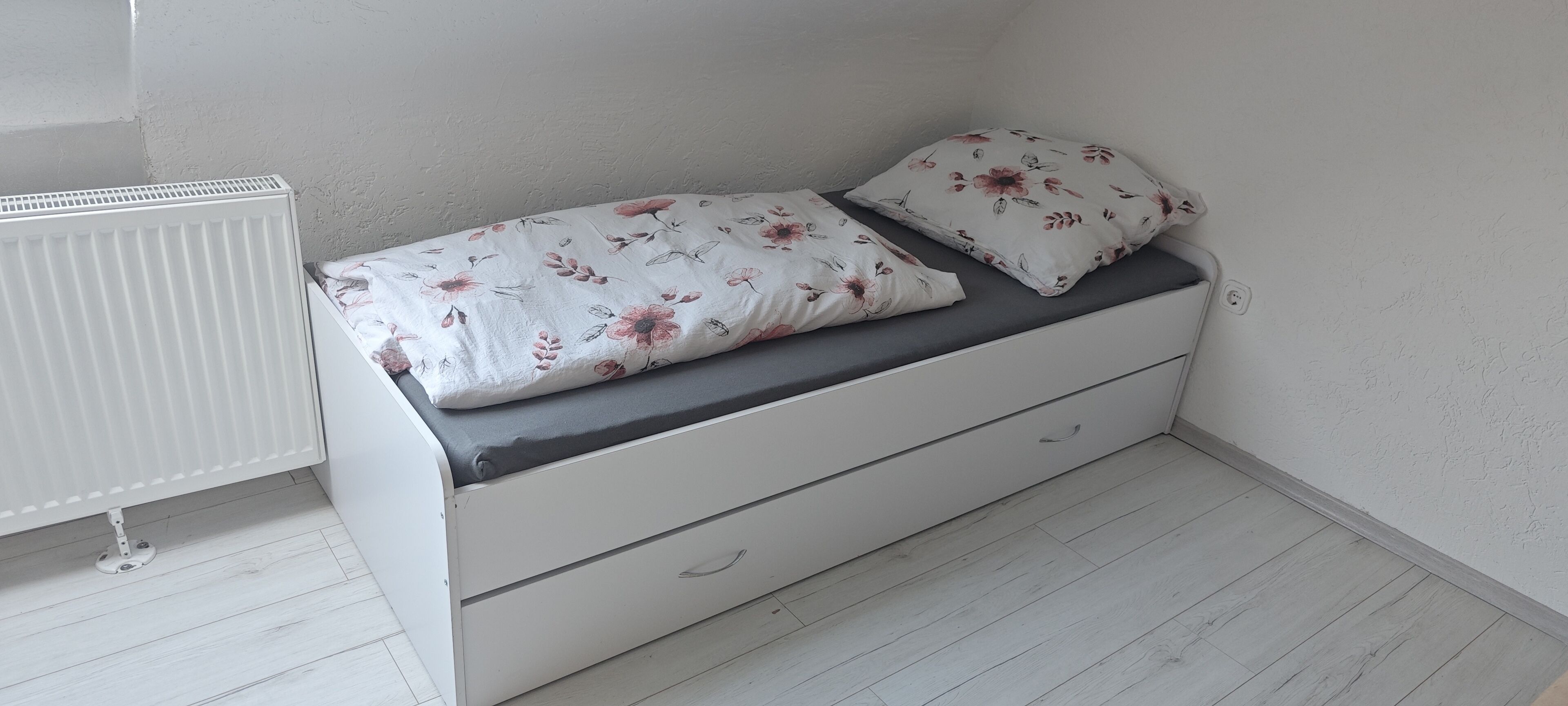 A small, white, single-bed room with a grey mattress and floral bedding.