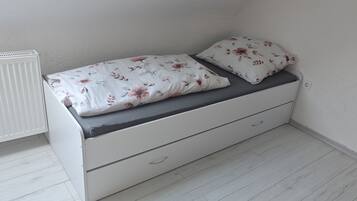A small, white, single-bed room with a grey mattress and floral bedding.