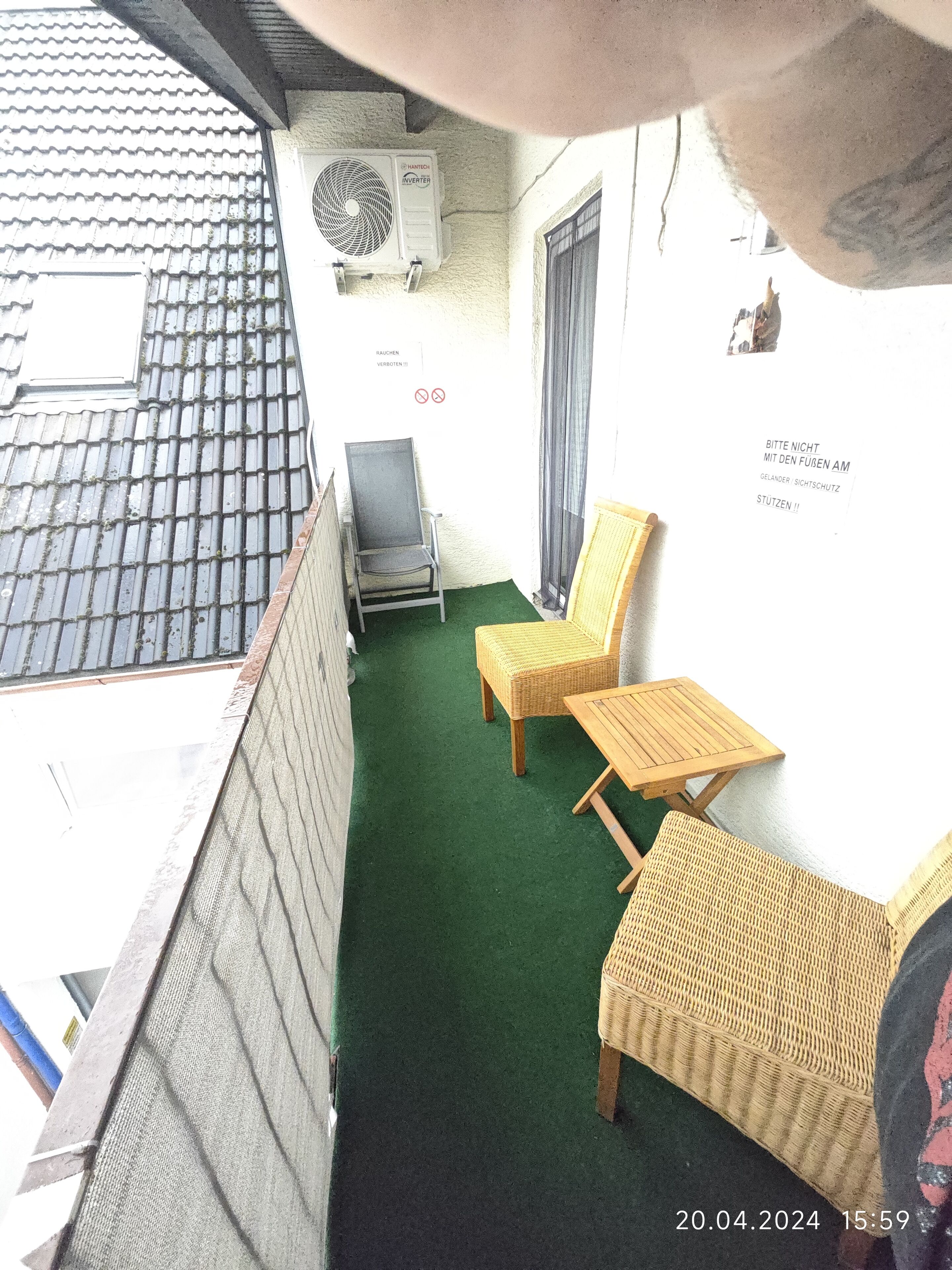 Een balkon met een vloer van groen kunstgras, twee rieten stoelen, een klein houten tafeltje en een plafondventilator.