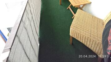 Een balkon met een vloer van groen kunstgras, twee rieten stoelen, een klein houten tafeltje en een plafondventilator.