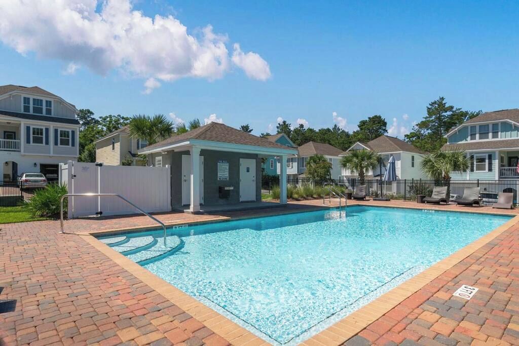 A swimming pool with a brick border, surrounded by houses with a covered pavilion, and a clear sky.