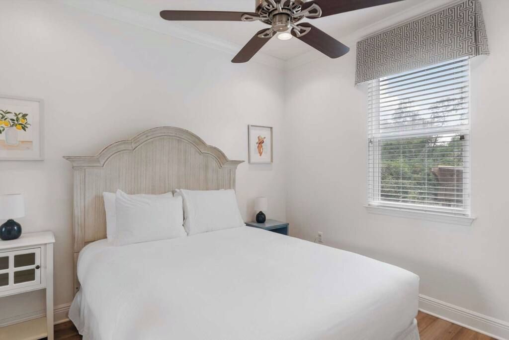 A bedroom with a bed, a nightstand, a ceiling fan, a window with blinds, and wall art.