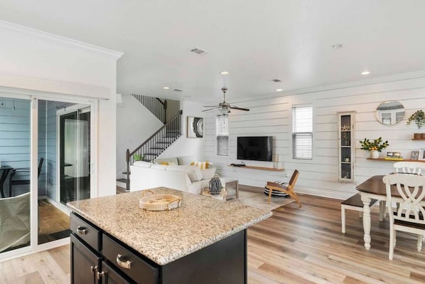 A modern kitchen with a granite countertop, a dining area with a table and chairs, a staircase leading to a living room, and a TV mounted on the wall.