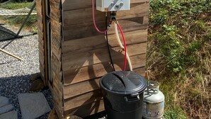 Ein tragbares Warmwassergerät mit Bedienfeld, verbunden mit einem schwarzen Eimer und einer Propangasflasche, vor einem Holzhintergrund mit Berglandschaft im Hintergrund.