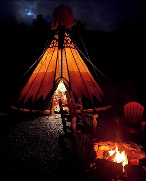 A tent with a ladder leading to a small interior space, a fire pit, and a chair outside.