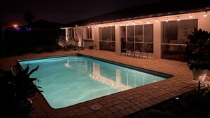Ein rechteckiger Swimmingpool mit einer gefliesten Kante, umgeben von einer Ziegelterrasse. In der Nähe eines überdachten Bereichs mit Lichterketten stehen Gartenstühle und ein Tisch.