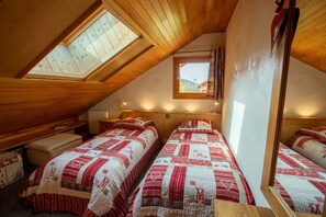 Ein gemütliches Schlafzimmer im Dachgeschoss mit zwei Betten, Holzbalken, einem Oberlicht und einem Fenster mit Blick auf ein Haus und Bäume.