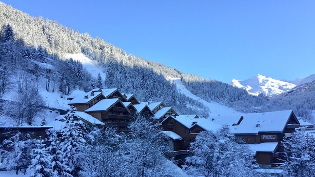 Eine schneebedeckte Lodge mit mehreren Holzhäusern, eine Skipiste im Hintergrund und eine gebirgige Landschaft.