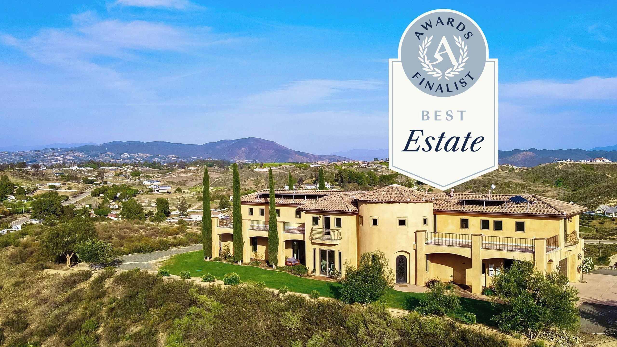 A large, multi-story building with a Mediterranean architectural style, surrounded by a well-maintained lawn and cypress trees, set against a backdrop of rolling hills and a clear sky.