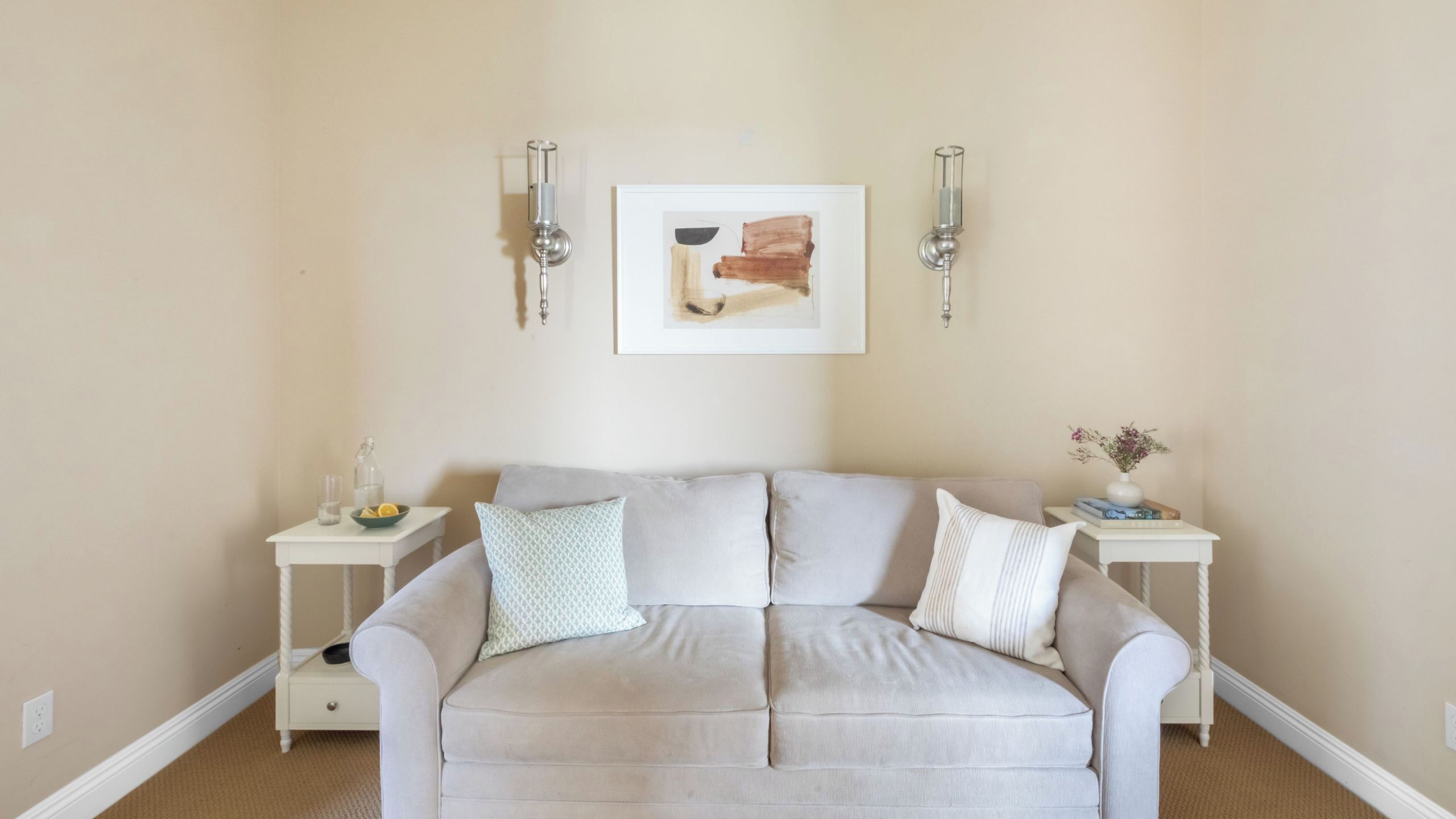 En stue i neutrale farver med en beige sofa, to sofaborde med pynteting og vægmonteret belysning.
