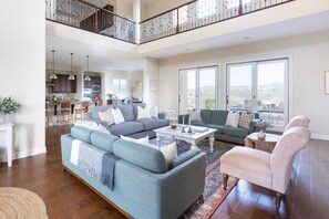 A spacious living room with a high ceiling, a balcony with a railing, and a view of the outdoors. - Palazzo Del Sol By Avantstay Breathtaking Home w/ Mountain Views & Hot Tub (Temecula)