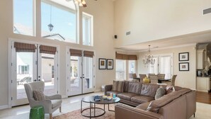 A spacious living room with a large sectional sofa, a round coffee table, and a comfortable armchair. There are large windows and a view of the outside.