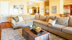 A living room with a sectional sofa, a coffee table with a bowl of fruit, and framed artwork on the walls. - Amarilla by Avantstay Peaceful Paradise w/ Pool, Spa & Theatre Room (La Quinta)